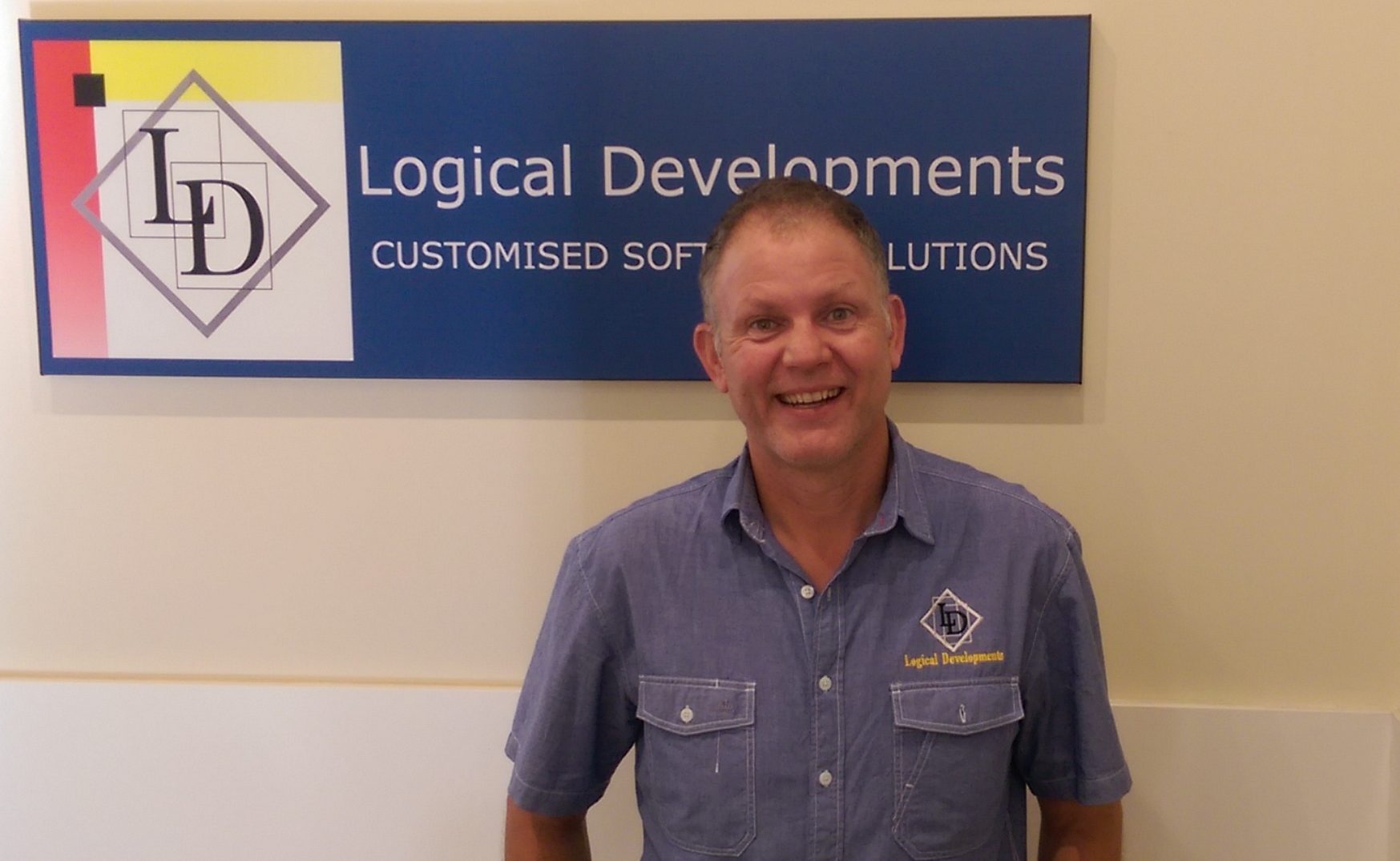 A picture of Shane. He stands beneath the Logical Developments banner that is hanging up in the stair case up the office. He wears the dress shirt that is part of the uniform and is smiling. 